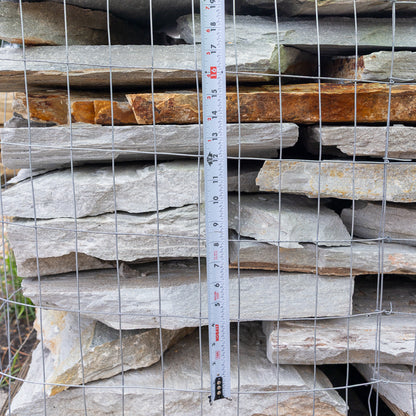 A stack of natural gray stone slabs behind a wire mesh fence, with a tape measure standing vertically to show the thickness of the slabs.